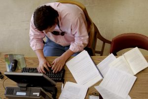 A photograph of a person sitting at a desk and typing on a computer. To the person's left is several notebooks.