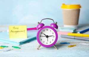 An image depicting time management. An analog clock, a sticky note with the words "time management" written on it, a cup of coffee, and various pens are visible.