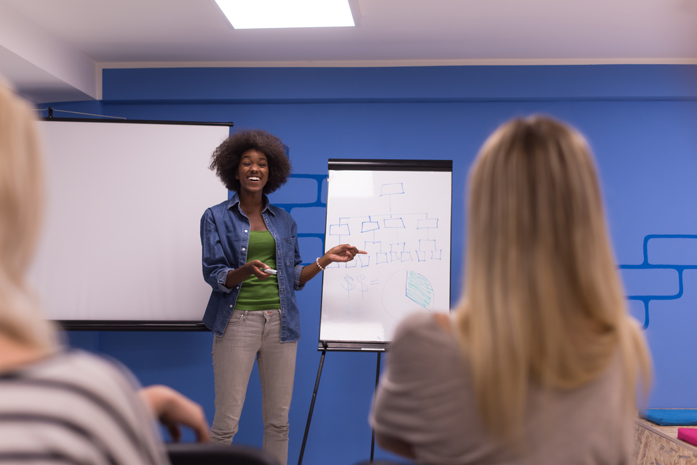 A woman standing at the front of a conference room, giving a presentation.