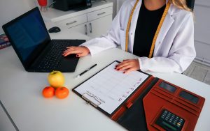A photo. A dietitian sits at her desk. She is typing on a laptop and has a patient's file open in front of her.