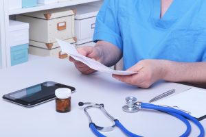 A photo. A pharmacist is sitting at a table. He is reviewing paperwork. There is a tablet device, a jar of pills, a stethoscope, and a clipboard on the table.
