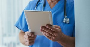 A closeup photo of a registered nurse. He is reading a tablet device that he is holding in his hands.
