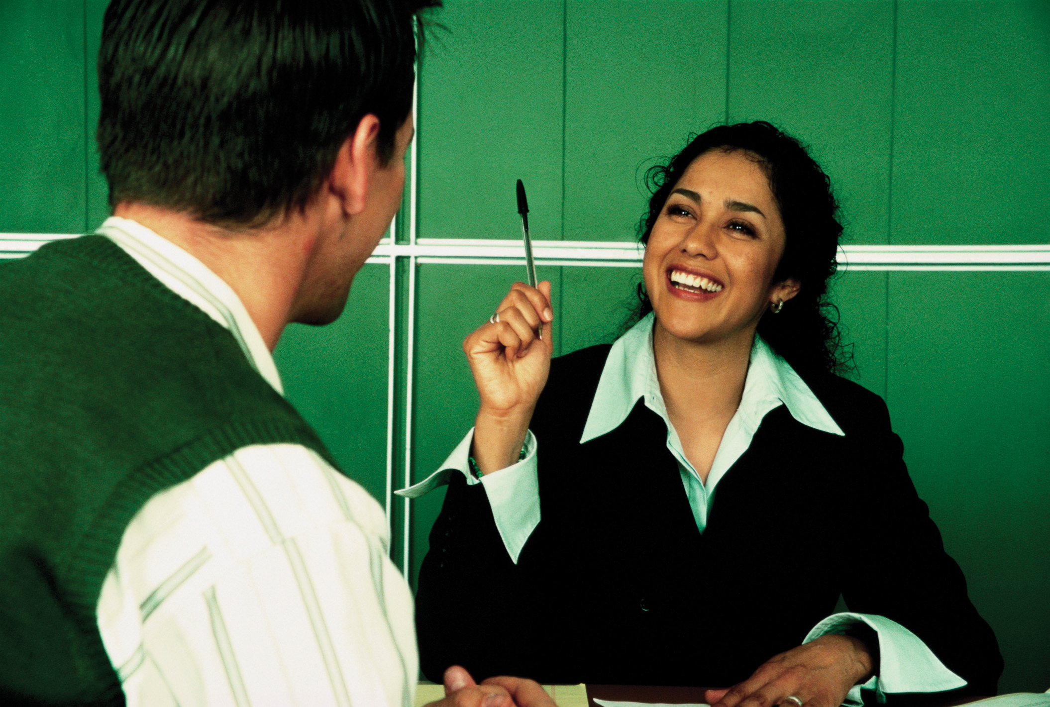A smiling woman gesturing with a pen, as she talks to a man across a desk.