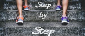 An image of sneaker clad feet walking up outdoor stairs with the words written in chalk on the steps, "Step by step."