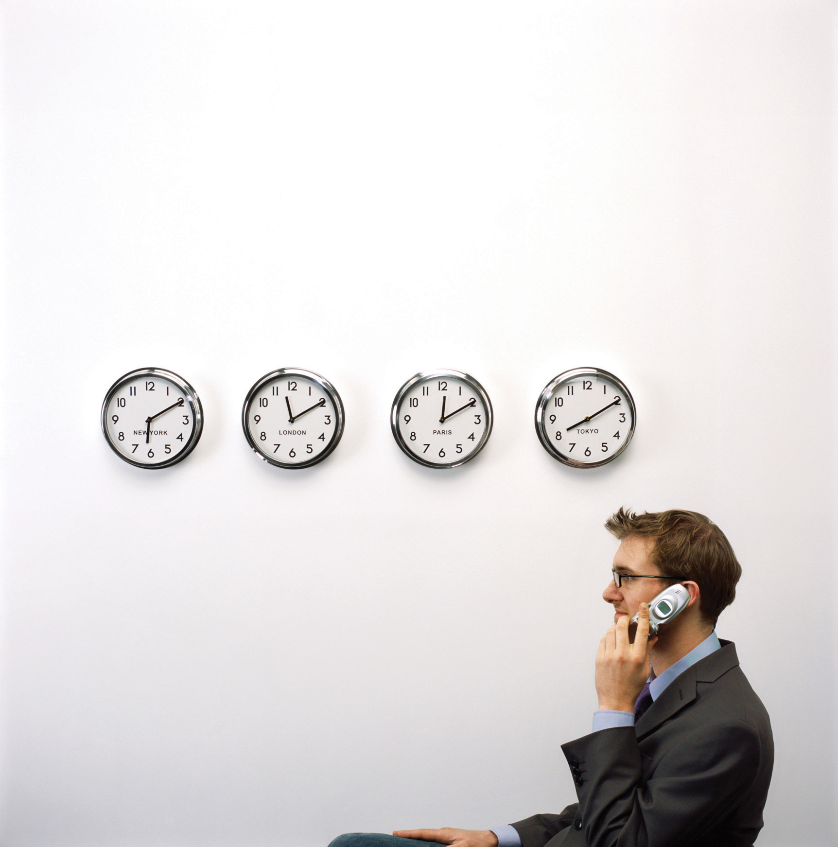 A man sits underneath a row of clocks from different time zones while he is on the phone.